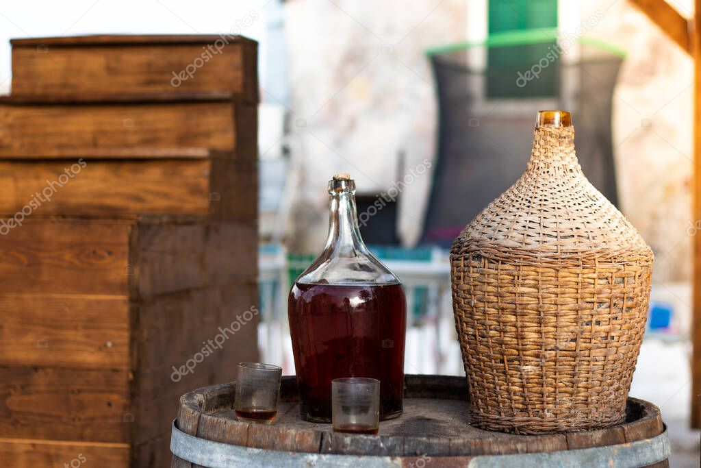 View of a glass bottle and a traditional wooden woven bottle holding several liters. Two empty glasses on the barrel. Small local meditarranean town, local domestic homemade wine