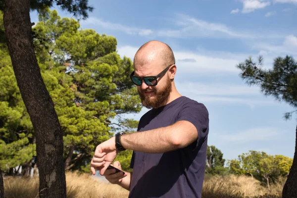 Young adult,bald and bearded wearing black sunglasses standing in nature on hot day checking his watch