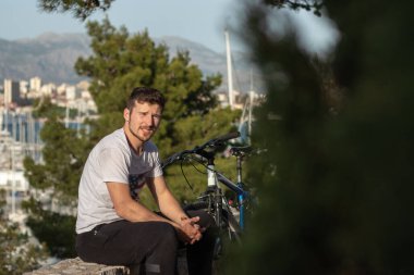 Çevresinde yeşil çimenler ve ağaçlar olan bisikletinin yanında dinlenen genç bir adam. Bisiklet sürücüsü sıcak bir günün tadını çıkarıyor.