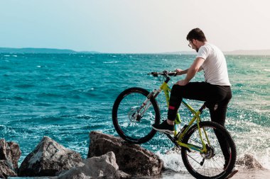 Hırvatistan 'ın Split kentinde, Mayıs 2020' de bir plaj kayasına yaslanmış bir bisikletle kıyıda duran genç adam. Rüzgârlı bir günde kıyıya vuran dalgalar onu ve çevreyi su baskınına uğratır.