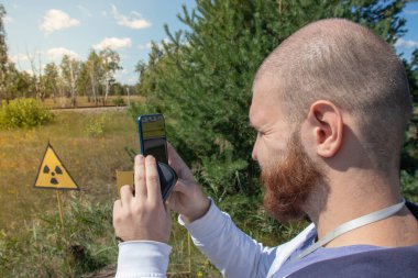 Çernobil sınırlama bölgesindeki yetişkin, kel bir turist elinde akıllı bir telefonla sarı uyarı işaretinin fotoğrafını çekiyor. Radyasyon bölgesini gösteriyor. Kızıl Orman 'ın önündeki bölge.
