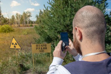 Çernobil sınırlama bölgesindeki yetişkin, kel bir turist elinde akıllı bir telefonla sarı uyarı işaretinin fotoğrafını çekiyor. Radyasyon bölgesini gösteriyor. Kızıl Orman 'ın önündeki bölge.