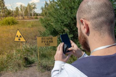 Çernobil sınırlama bölgesindeki yetişkin, kel bir turist elinde akıllı bir telefonla sarı uyarı işaretinin fotoğrafını çekiyor. Radyasyon bölgesini gösteriyor. Kızıl Orman 'ın önündeki bölge.