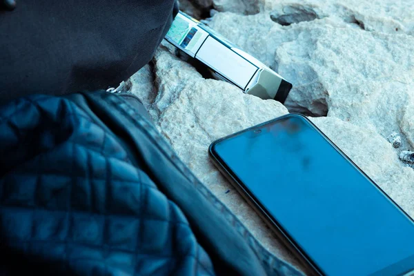 Smartphone laying on a rock with a pack of cigarettes behind it. Jacket and backpack next to it, early morning relaxation, hiking outisde