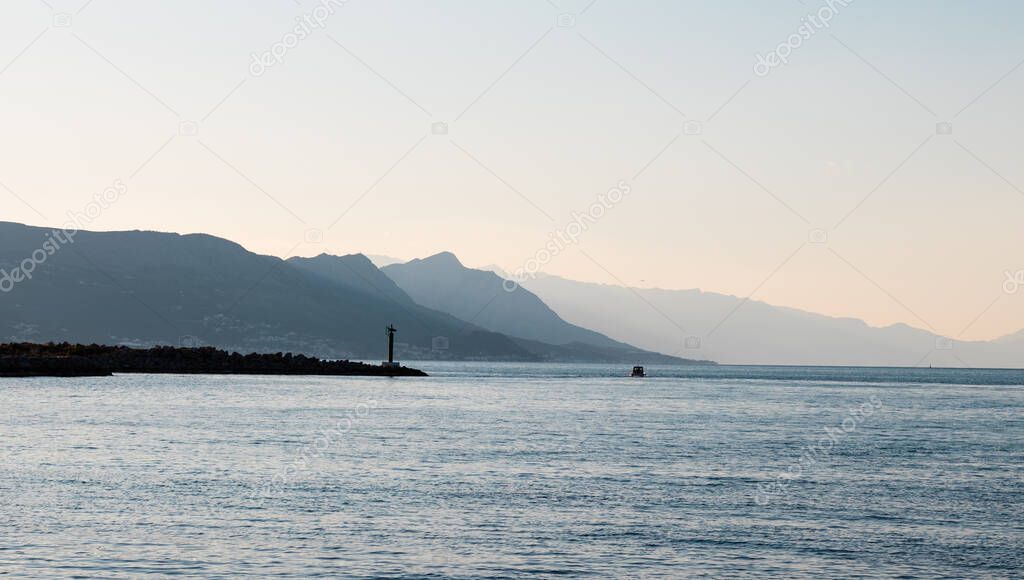 Bright blue morning sunrise on the shore of split, lighthouse in the distance with mountain silhouettes going into the distance. Small local boat on the sea