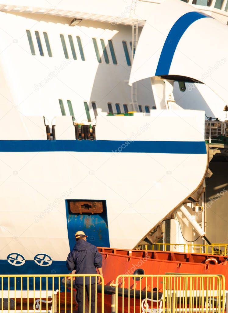 Ferry ship ocean liner worker standing behind a yellow fence, seen from a distance, vertical shot with the opened ferry visible behind him loading cars