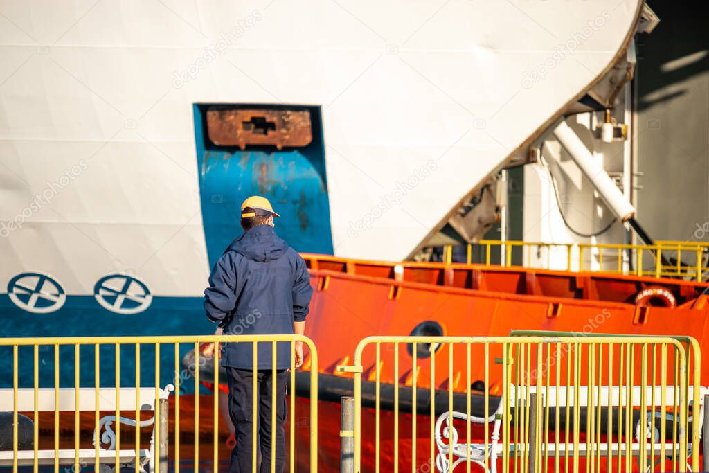 Ferry ship ocean liner worker standing behind a yellow fence, seen from a distance, multiple huge ships in the distance. Part of a white ferry and orange tugboat
