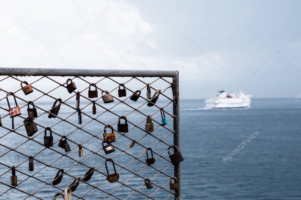 Rusted padlocks and fence on a rainy day drenched in water near the sea. Blurred ferry passing by in the distance. Dark day, broken love and negative emotions