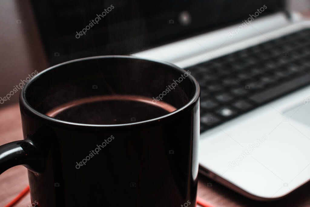 Black coffee mug with a dark coffee inside steaming, silver laptop in the background. Concept of programming , graphic design, online work