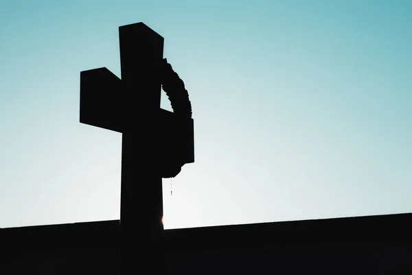 Black outline of a christan cross with a tiny one hanging from it. Contrast of black underexposed cross and bright teal blue morning sky
