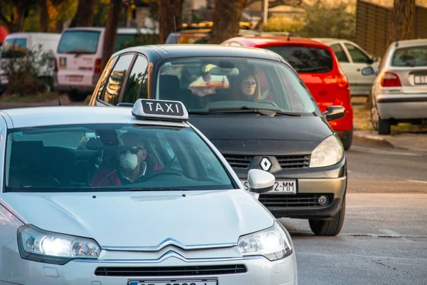 Corona virüsü salgını sırasında koruyucu maske takan beyaz bir Citroen 'in içindeki taksi şoförü. Uzakta endişeli bir kadın var.