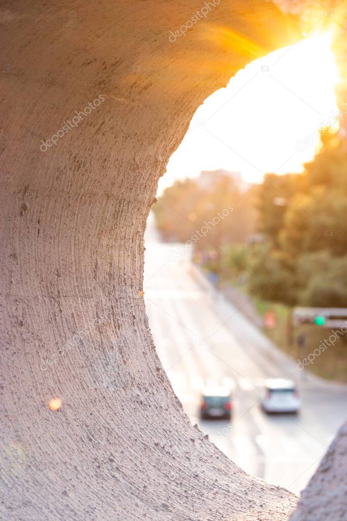 Wall of a bridge in focus, road underneath with multiple cars out of focus passing by. Busy road, green light on the intersection. Sunny day