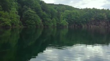 Hırvat ulusal parkı Plitvice göllerindeki büyük bir gölün üzerinde ağır çekim dönüyor. Orman su yüzeyini yansıtıyor, günün sonunda güneş batıyor.