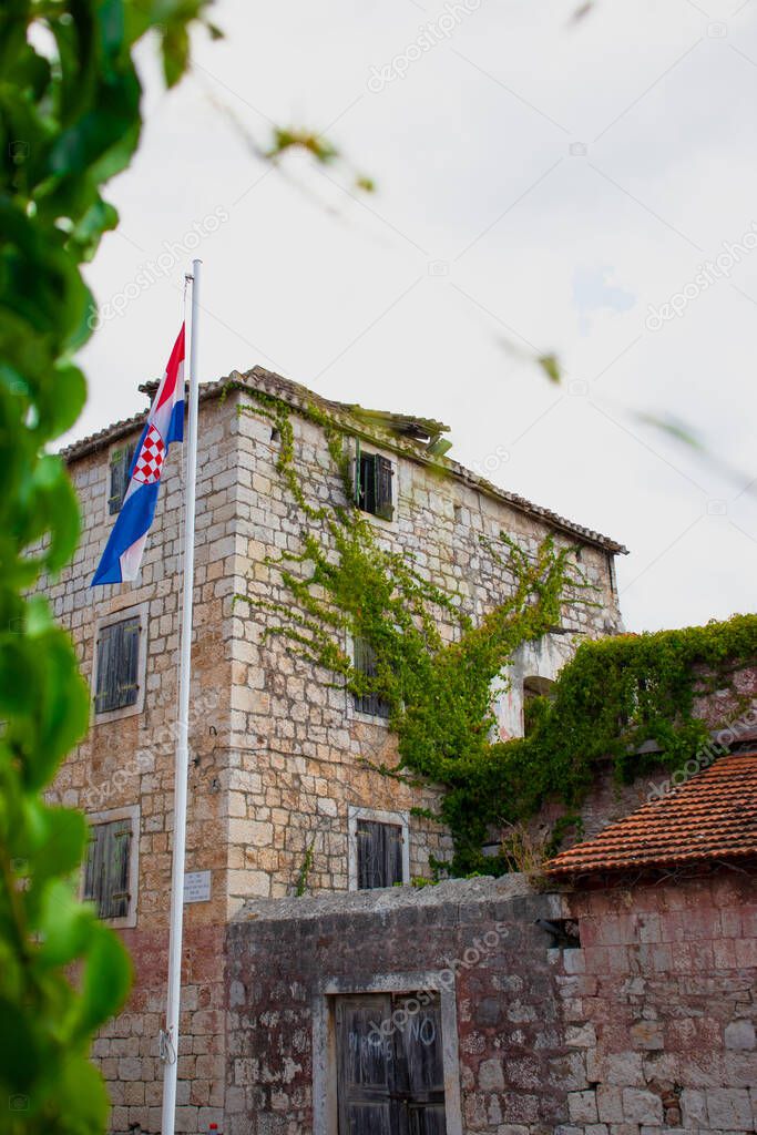 Beautiful old stone house in the village of Milna overgrown with green plants, croatian flag on a pole waving in front of it