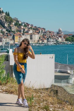 Sıcak bir yaz gününde, deniz kenarında poz veren mavi etekli ve turuncu bluzlu çekici bir esmer. Rüzgarda uçuşan uzun saçlar. Uzakta Sibenik kasabasının ana hatları. Nüfuzlu kişiler seyahat eder