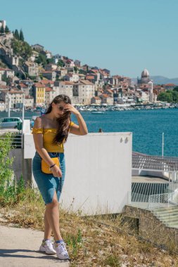 Sıcak bir yaz gününde, deniz kenarında poz veren mavi etekli ve turuncu bluzlu çekici bir esmer. Rüzgarda uçuşan uzun saçlar. Uzakta Sibenik kasabasının ana hatları. Nüfuzlu kişiler seyahat eder
