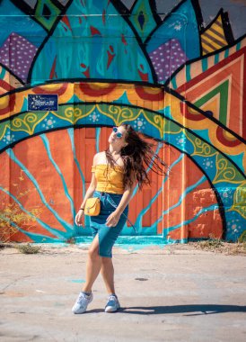 Çekici esmer, mavi etekli ve turuncu bluzlu bir kız yaz gününde arka planda renkli bir duvarla poz veriyor. Sarı çanta, şık etken seyahat ve modellik.