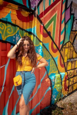Çekici esmer, mavi etekli ve turuncu bluzlu bir kız yaz gününde arka planda renkli bir duvarla poz veriyor. Sarı çanta, şık etken seyahat ve modellik.