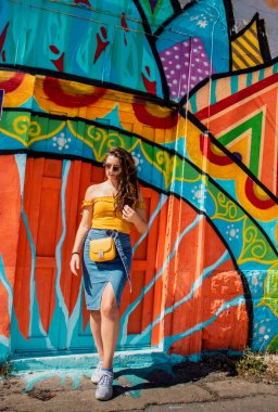 Çekici esmer, mavi etekli ve turuncu bluzlu bir kız yaz gününde arka planda renkli bir duvarla poz veriyor. Sarı çanta, şık etken seyahat ve modellik.