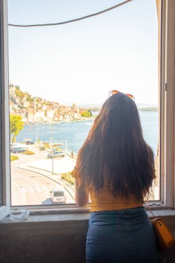 Esmer, uzun saçlı, arkadan görünen, bir pencere çerçevesinde duran, uzaklığa bakan. Uzakta Sibenik kasabasının ana hatları. Hırvatistan ve Adriyatik Denizi 'ni gezmek