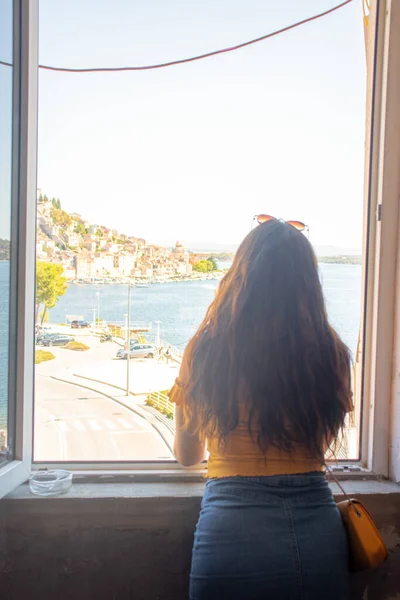 Esmer, uzun saçlı, arkadan görünen, bir pencere çerçevesinde duran, uzaklığa bakan. Uzakta Sibenik kasabasının ana hatları. Hırvatistan ve Adriyatik Denizi 'ni gezmek