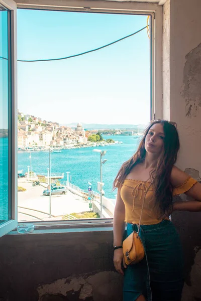 Attractive brunette posing and smiling near a window, coastal town of sibenik, croatia in the distance. Traveling Croatia and the adriatic sea, positive vibes during the summer