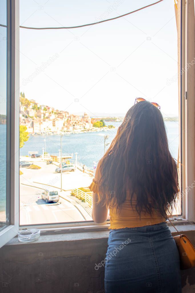 Brunette with long hair seen from behind, standing on a window frame looking into the distance. Outline of the town of Sibenik in the distance. Traveling Croatia and the adriatic sea