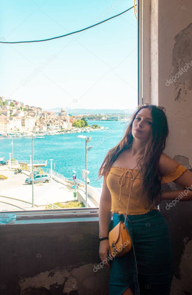 Attractive brunette posing and smiling near a window, coastal town of sibenik, croatia in the distance. Traveling Croatia and the adriatic sea, positive vibes during the summer