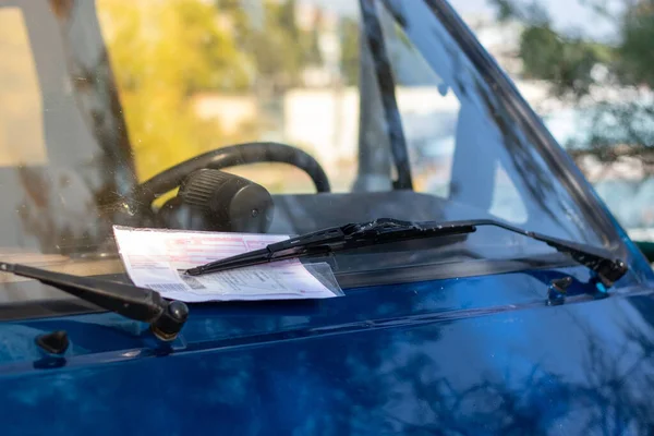 Red parking ticket stuck under the windshielf wiper of a blue car, person fined for illegal parking in public. Contents of the ticket blurred for privacy