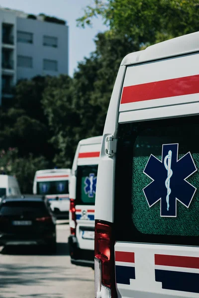 Hırvatistan 'ın Split kentinde Coronavirus salgını sırasında park etmiş beyaz bir ambulansın arka tarafındaki dikey çekim. Birden fazla minibüs sıraya dizilmiş, kapılarında sağlık sembolleri var.