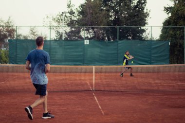 Mavi gömlekli ve mavi pantolonlu bir adam tenis oynarken görülmüş, geniş açık turuncu kil oyun alanı. Rakibin topa vuruşunun ana hatlarıyla diğer taraftan servis yapmayı bekliyor.