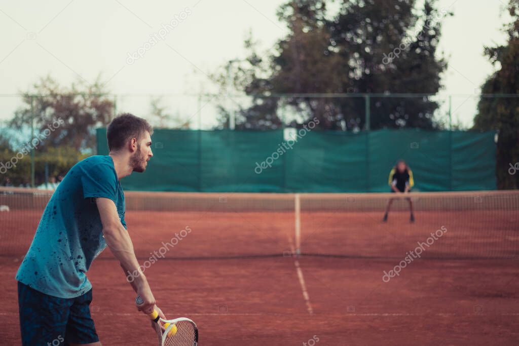 Back view of a man holding a racket and a ball, prepairing to serve while playing a match of tennis outside on an orange clay field
