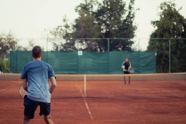 Mavi gömlekli ve mavi pantolonlu bir adam tenis oynarken görülmüş, geniş açık turuncu kil oyun alanı. Rakibin topa vuruşunun ana hatlarıyla diğer taraftan servis yapmayı bekliyor.