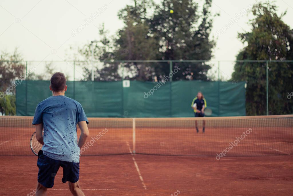 Man in blue shirt and blue pants seen from behind playing tennis, wide open clay orange playing field. Anticipating the serve from the other side, outline of the opponent hitting a ball