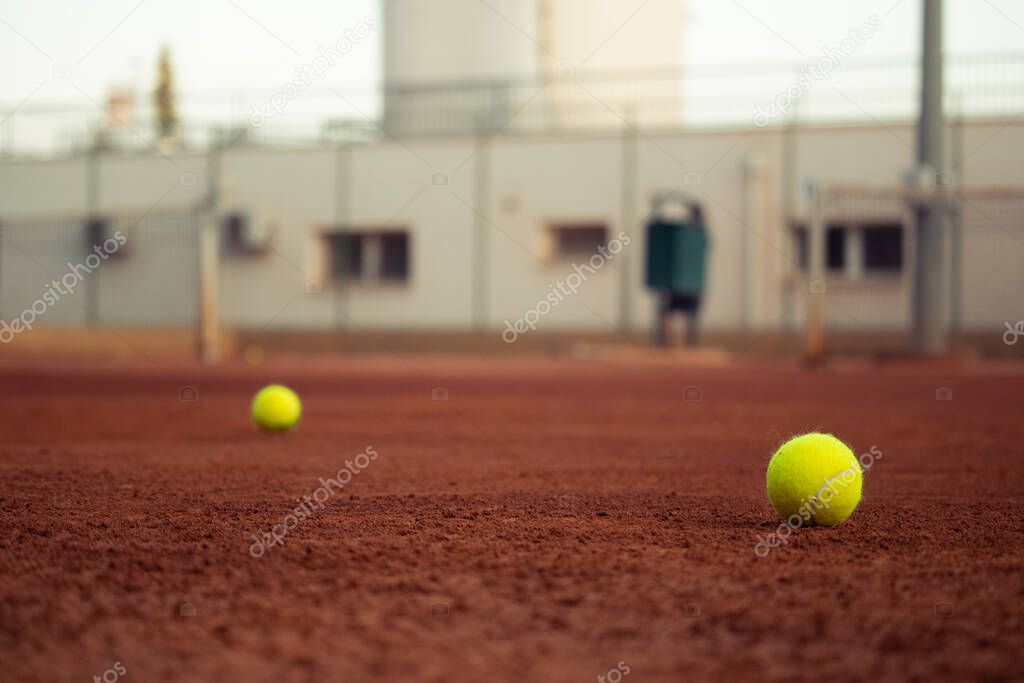 Two yellow tennis balls standing in the orange ground clay playing field, one further than the other. Concepts of playing tennis outside on clay