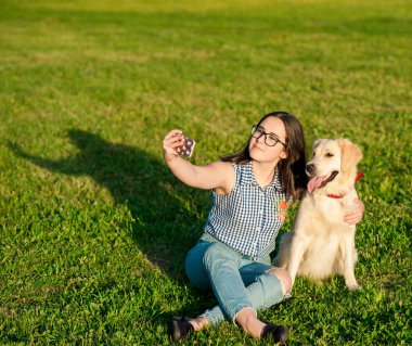 Yeşil çayırda Golden Retriever köpeğiyle selfie çeken neşeli, genç bir kadın. Güzel kız ve köpeği telefonla kendi portresini yapıyorlar.