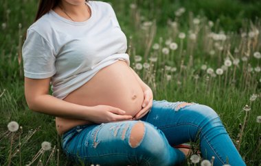 Genç bir kadın elleriyle karnına dokunur ve kalp şekli oluşturur. Hamilelik, aşk, insanlar ve beklenti konsepti