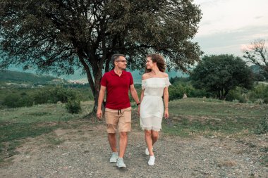 Genç çift dağlarda el ele tutuşup birbirlerine bakarak yürüyor.