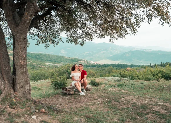 Joven pareja feliz se sienta en un salto cerca de un hermoso árbol en el telón de fondo de las montañas —  Fotos de Stock