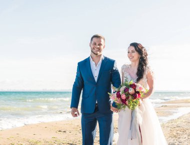 Güzel bir düğün çifti, damat ve gelin, deniz kıyısında yürüyor, koşuyor, gülüyor, eğleniyorlar. Düğün istirahatı, rahatlama balayı konsepti.