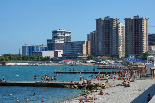 Városi strand és hullámtörők a modern apartmanházak hátterében. Az emberek fürdenek és napoznak.. — Stock Fotó