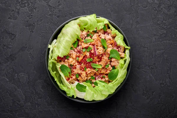 Laab Gai - Thai Minced Chicken Salad in black bowl at dark slate background. Larb is thailand cuisine dish with minced meat, fish sauce, lime juice, mint, lettuce and roasted rice. Thai Food.