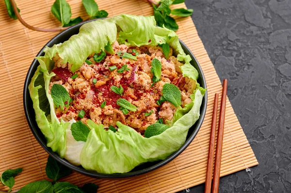 Laab Gai - Thai Minced Chicken Salad in black bowl at dark slate background. Larb is thailand cuisine dish with minced meat, fish sauce, lime juice, mint, lettuce and roasted rice. Thai Food