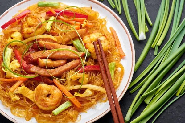 Singapore Mei Fun in white plate on dark slate background. Singapore Noodles is chinese cuisine dish with rice noodles, prawns, char siu pork, carrot, red onion, napa cabbage. Chinese food.