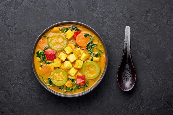 A Yellow Veg Thai Curry with Tofu and vegetables in black bowl at dark slate background. Vegetarian Thai Curry with tofu, zucchini, bell pepper, spinach, carrot. Thai Food. Indian vegetable curry