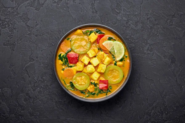 Yellow Veg Thai Curry with Tofu and vegetables in black bowl at dark slate background. Vegetarian Thai Curry with tofu, zucchini, pepper, spinach, carrot. Thai Food. Indian vegetable curry. Copy space