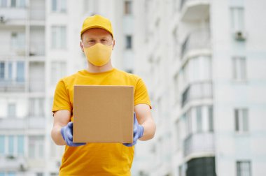 Teslimatçı eleman sarı üniforma şapkası, tişört, yüz maskesi ve eldiven içinde bina zemininde karton bir paket tutuyor. Covid-19 virüs salgınında güvenlik karantina hizmeti.
