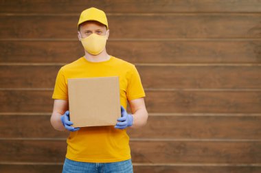 Teslimatçı eleman sarı üniforma şapkası, tişört, yüz maskesi ve eldivenler içinde ahşap arka planda bir karton kutu tutuyor. Covid-19 virüs salgınında güvenlik karantina hizmeti.