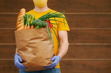 Sarı üniforma şapkalı, tişörtlü, yüz maskeli, eldivenli market teslimatçısı içinde yiyecek, meyve ve sebze bulunan bir kese kağıdı tutuyor. Covid-19 virüs salgınında karantina dağıtım hizmeti.