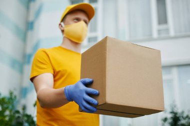 Teslimatçı adam sarı üniforma şapkası, tişört, yüz maskesi ve eldiven içinde bina zemininde karton bir paket tutuyor. Covid-19 virüs salgınında güvenlik karantina hizmeti.